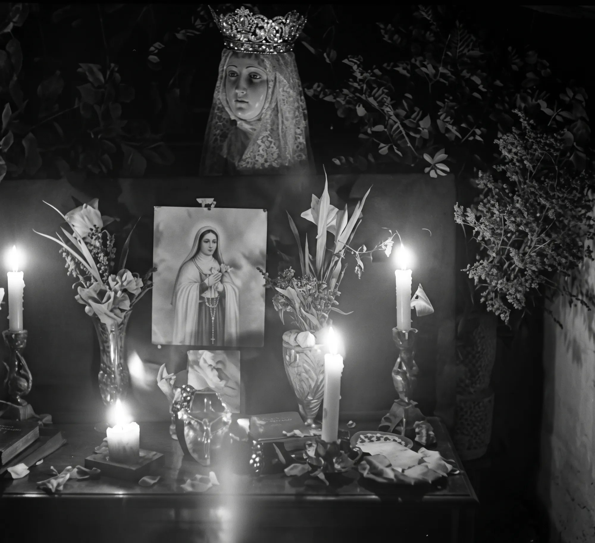 A candlelit altar: a crowned and veiled figure above flowers, candlesticks and a framed devotional image.